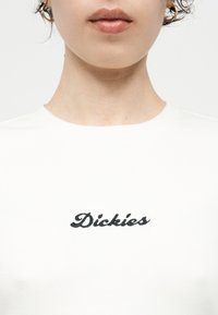 Κοντινό πλάνο του κάτω μέρους του προσώπου και του λαιμού ενός ατόμου που φοράει λευκό πουκάμισο με την επιγραφή «Dickies» κεντημένη με μαύρο χρώμα στο στήθος.