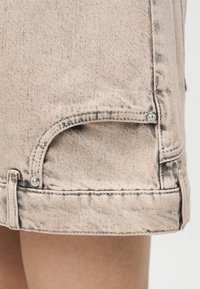 Κοντινό πλάνο στο τελείωμα μπεζ τζιν φούστας με μεταλλικά καρφιά και εμφανείς ραφές πάνω από γυμνό πόδι.