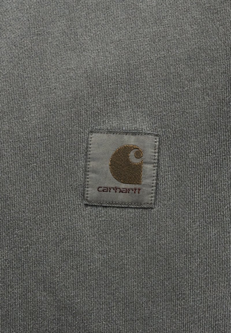 Gray knitted fabric with a square Carhartt logo patch featuring a brown crescent shape and text in dark red.