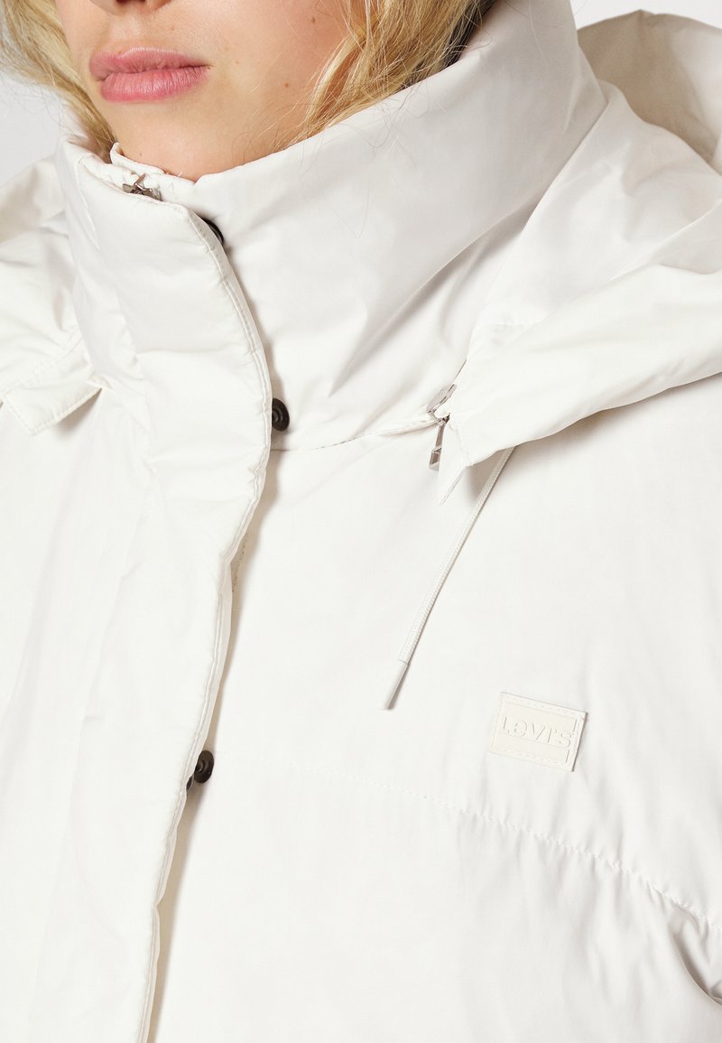 Chaqueta de plumas blanca con un cuello alto, que cuenta con una tela texturizada, detalles de botones y un pequeño parche con logo en el lateral inferior.