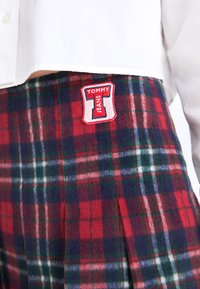 Red and green plaid skirt made of soft fabric, featuring a pleated design and a prominent embroidered "TOMMY JEANS" logo patch.
