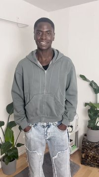 Fleece cinza com fecho, adornado com detalhes de costura texturizada, combinado com jeans claros desgastados com bordas desfiadas. Plantas ao fundo.