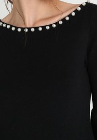 Czarny sweter z prążkowanej tkaniny, z szerokim dekoltem zdobionym rzędem dekoracyjnych pereł. Miękka faktura, dopasowany fason.