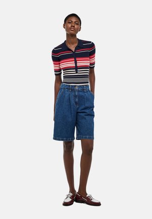 Femme debout, mains dans les poches, portant un polo à manches courtes rayé bleu marine, rouge et blanc, ainsi qu'un short en jean ample arrivant aux genoux.