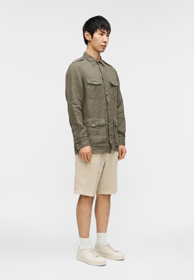 Πουκάμισο από λινό ελιάς με δύο μπροστινές τσέπες και κούμπωμα με κουμπιά, σετ με σορτς μπεζ από λινό και λευκά αθλητικά παπούτσια. Απλός και λειτουργικός στιλ.