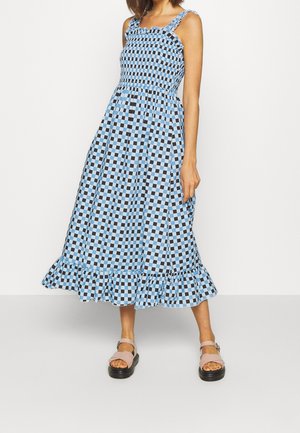Femme portant une robe midi sans manches à carreaux bleu et noir avec un ourlet à volants et des sandales compensées beige, debout devant un fond blanc.