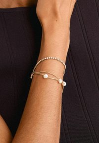 Pulseras doradas en un brazo: una con cuentas transparentes, otra con perlas blancas, apiladas para un aspecto texturizado sobre un fondo oscuro.