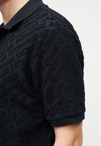 Κοντινή λήψη μαύρου πόλο με υφασμένο, ανάγλυφο κείμενο που επαναλαμβάνει τη φράση «ARMANI EXCHANGE» σε όλο το ύφασμα.