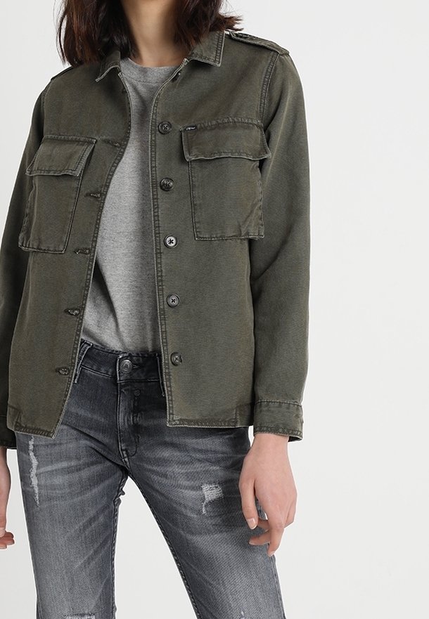 Personne portant une veste verte à boutons avec deux poches poitrine sur un t-shirt gris et un jean gris usé.