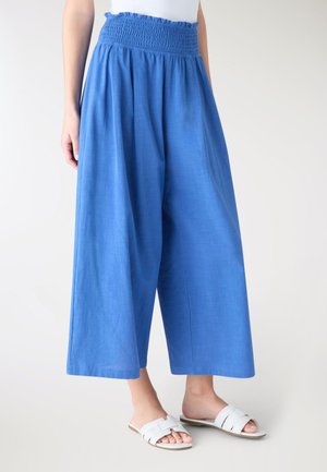 Donna che indossa pantaloni blu a vita alta e gamba larga con vita elastica arricciata e sandali bianchi slip-on, in piedi su uno sfondo neutro.