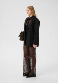 Svart skräddarsydd blazer med två knappar, lagd över en genomskinlig spetsklänning med blommönster. Modellen håller en olivgrön väska.