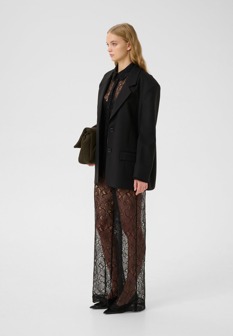 Schwarzer taillierter Blazer mit zwei Knöpfen, über einem durchsichtigen Spitzenkleid mit floralem Muster getragen. Das Model hält eine olivgrüne Tasche.