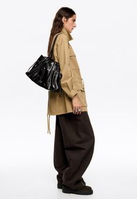 Beżowa kurtka robocza, czarna torebka z lakierowanej skóry, oversize'owe brązowe spodnie, teksturowana tkanina, warstwowy design, srebrne pierścionki.