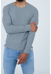 Maglione grigio a maglia con scollatura rotonda, maniche aderenti e texture a coste; abbinato a jeans blu chiaro. Etichetta visibile all'orlo.