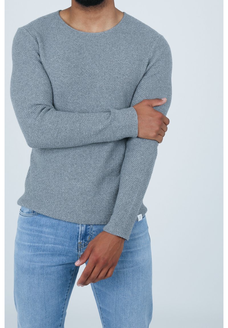 Maglione grigio a maglia con scollatura rotonda, maniche aderenti e texture a coste; abbinato a jeans blu chiaro. Etichetta visibile all'orlo.