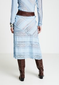 Robe en dentelle bleu clair à manches longues et design en couches, avec une ceinture en cuir marron cloutée. Portée avec des bottes marron hautes jusqu'aux genoux.