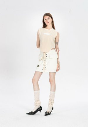 Joven mujer de pie vistiendo una blusa sin mangas beige, falda blanca con cordones y bolsillos, calentadores de piernas beige arrugados y zapatos negros de tacón.