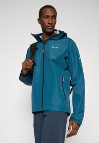 Τιρκουάζ αδιάβροχο μπουφάν με μπροστινό φερμουάρ, ρυθμιζόμενη κουκούλα και πορτοκαλί λεπτομέρειες. Διαθέτει κομψό σχέδιο και διακριτική σήμανση.