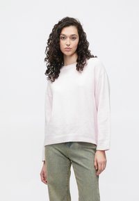 Svetlo roza, teksturiran pulover z okroglim izrezom in spuščenimi rameni, kombiniran z obledelimi zelenimi kavbojkami. Preprost, priložnostni dizajn.