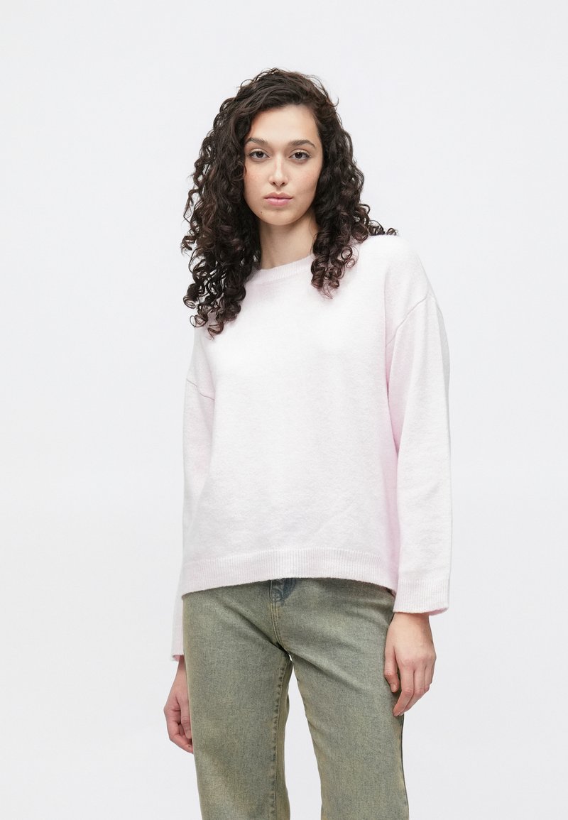 Svetlo roza, teksturiran pulover z okroglim izrezom in spuščenimi rameni, kombiniran z obledelimi zelenimi kavbojkami. Preprost, priložnostni dizajn.