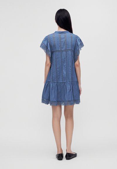 Femme aux longs cheveux foncés, vue de dos, portant une robe bleue à manches courtes avec des détails en dentelle et des chaussures plates noires sur fond uni.