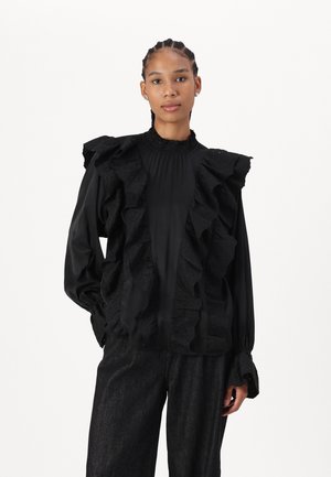 Blusa negra con detalles de volantes en la parte delantera y los hombros, con cuello alto y mangas largas, combinada con pantalones oscuros.