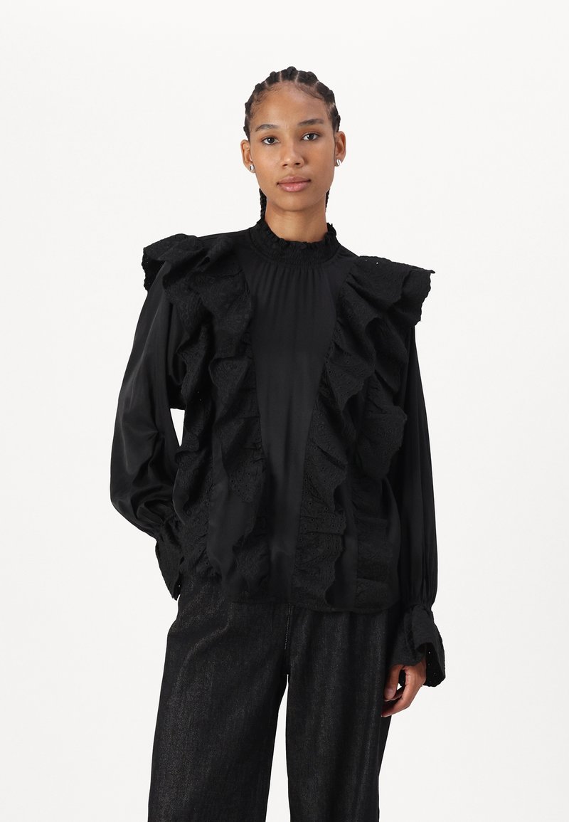 Blusa negra con detalles de volantes en la parte delantera y los hombros, con cuello alto y mangas largas, combinada con pantalones oscuros.