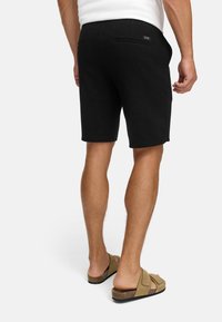 Hombre que lleva pantalones cortos negros, camisa blanca y sandalias beiges, mostrado de espaldas contra un fondo blanco liso.