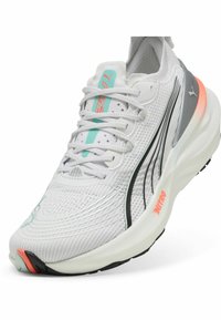 Zapatilla deportiva blanca con parte superior de malla, acentos gris claro, suela negra y detalle coral en el talón. Presenta cordones texturizados y un logo.