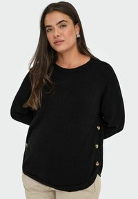 Donna con lunghi capelli scuri indossa un maglione nero con bottoni laterali e pantaloni beige, in piedi davanti a uno sfondo chiaro e uniforme.