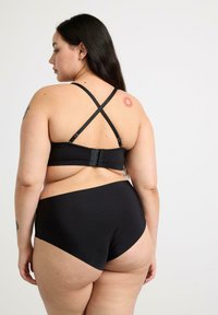 Soutien-gorge noir avec des bretelles croisées au dos et une fermeture à crochet. Culotte haute sans coutures en noir. Le modèle montre un tatouage de soleil sur l'épaule.
