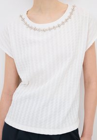 Top blanco de manga corta con un patrón texturizado, cuello redondo adornado con cuentas plateadas en forma de flor y un ajuste holgado.