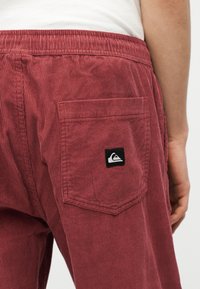 Κοντινό πλάνο της πίσω τσέπης κόκκινων βελούδινων παντελονιών με το λογότυπο της Quiksilver στην περιοχή του ισχίου ενός ατόμου και μέρος του χεριού του ορατό.