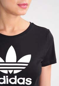 Svart bomulls-t-shirt med rund halsringning, korta ärmar och en framträdande vit Adidas trefoil-logotyp med horisontella ränder.