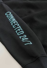 Schwarzer Sweatshirt mit strukturiertem Finish, ausgestattet mit einem hellblauen "CONNECTED 24/7" Grafik auf dem linken Ärmel. Rippbündchen sind sichtbar.