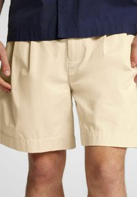 Pantaloni corti beige realizzati in tessuto leggero con una vestibilità ampia, dotati di orlo arrotolato e tasche laterali. Abbinati a una camicia blu scuro.