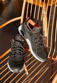 Paire de chaussures de sport noires avec des semelles blanches et une doublure orange, posée sur une chaise en métal orange à la lumière du soleil.