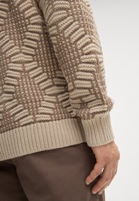 Strickpullover mit einem strukturierten Muster in Beige und Braun, mit gerippten Bündchen und Saum. Das Material wirkt weich und hat eine grobe Webart.