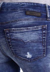 Tmavě modré džíny z denimu s ošoupanou zadní kapsou a černou koženou nášivkou. Obsahují mírné vyblednutí a kontrastní prošití. Klasický střih.