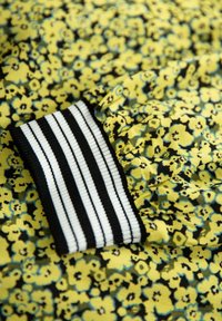 Musterstoff mit gelbem Blumenmuster auf schwarzem Hintergrund. Verfügt über einen schwarz-weißen gestreiften Rippbund. Weiche Textur.