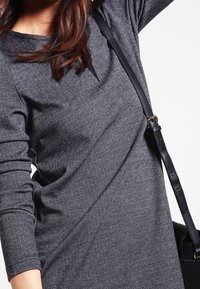 Długa sukienka w szarym wzorze w jodełkę z długimi rękawami, wykonana z teksturowanej tkaniny, charakteryzująca się dopasowanym krojem z ukośnym szwem oraz czarną torebką na ramię.