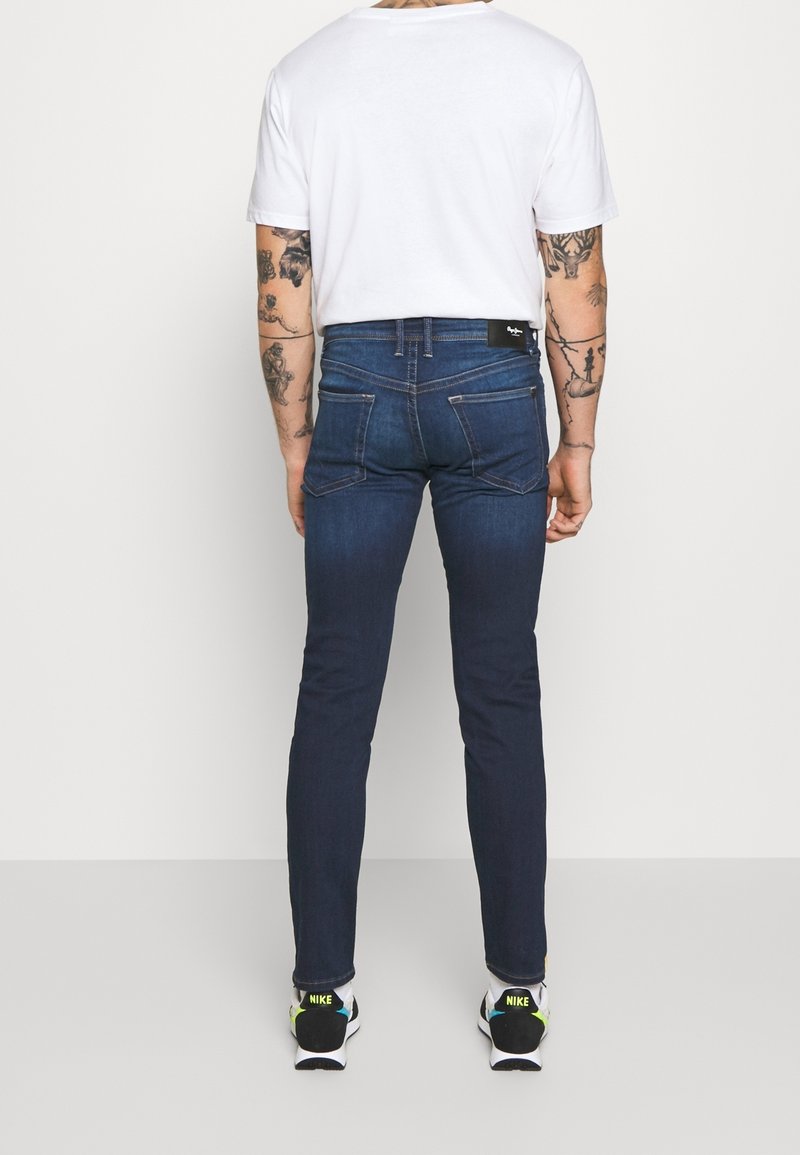 Homme aux bras tatoués portant un jean slim bleu foncé, un t-shirt blanc et des baskets blanches Nike, se tenant devant un fond blanc.