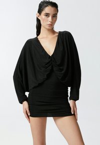 Abito nero realizzato in tessuto lucido e elasticizzato. Presenta un profondo scollo a V, una parte anteriore drappeggiata e maniche lunghe con dettagli arricciati.