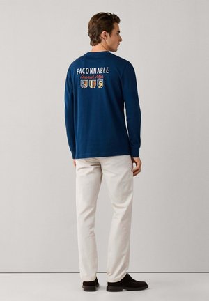 Hombre de pie de espaldas, con una camisa azul marino de manga larga con los logos "Façonnable French Alps" y escudo en la espalda, pantalones beige y zapatos negros.
