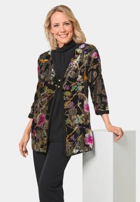 Blumenmuster-Cardigan mit schwarzem Hintergrund, der lebendige Farben, 3/4-Ärmel und dekorative Knöpfe aufweist, über einem schwarzen Oberteil getragen.