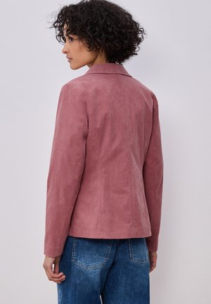 Žena s kudrnatými vlasy, oblečená v růžovém saku a modrých džínách, stojící bokem před jednoduchým světlým pozadím.