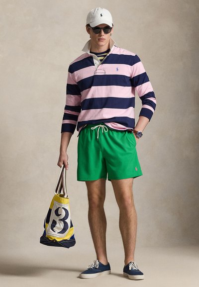Πολό μακρυμάνικο με ρίγες σε ροζ και ναυτικό μπλε, πράσινα σορτς για κολύμβηση, ναυτικά παπούτσια τύπου slip-on και τσάντα τύπου tote σε κίτρινο και ναυτικό μπλε με λογότυπο. Άσπρο καπέλο και γυαλιά ηλίου.