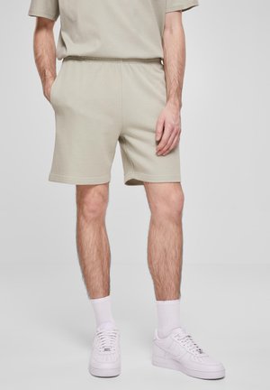 Man wearing beige shorts and matching t-shirt with white socks and white sneakers, standing with one hand in pocket.