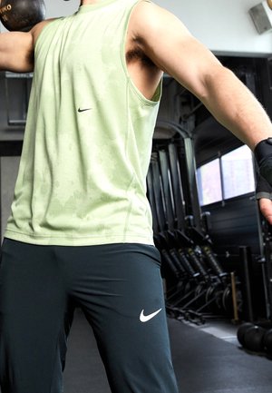 Homme soulevant une kettlebell les bras tendus dans une salle de sport, portant un débardeur et des gants, équipement de gym visible en arrière-plan.