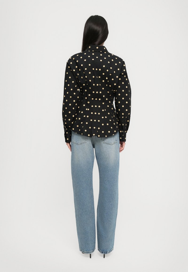 Vrouw die met haar rug naar de camera staat, gekleed in een zwart shirt met beige stippen en lichte spijkerbroek met rechte pijpen tegen een witte achtergrond.